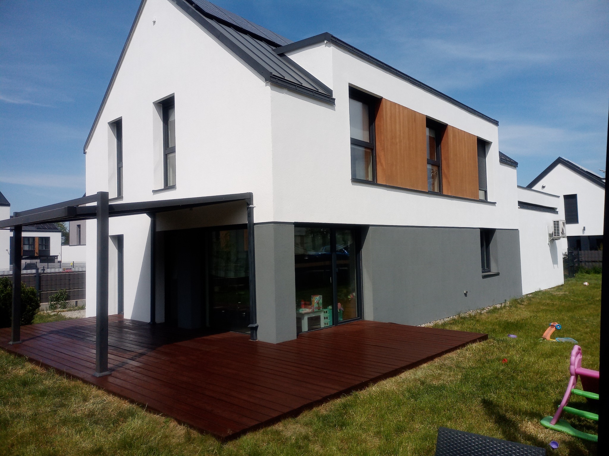 Nowoczesny dom jednorodzinny z białą elewacją, szarą podmurówką, drewnianymi panelami, ciemną stolarką okienną i panelami słonecznymi na dachu, widoczny z perspektywy tarasu z ciemnego drewna.