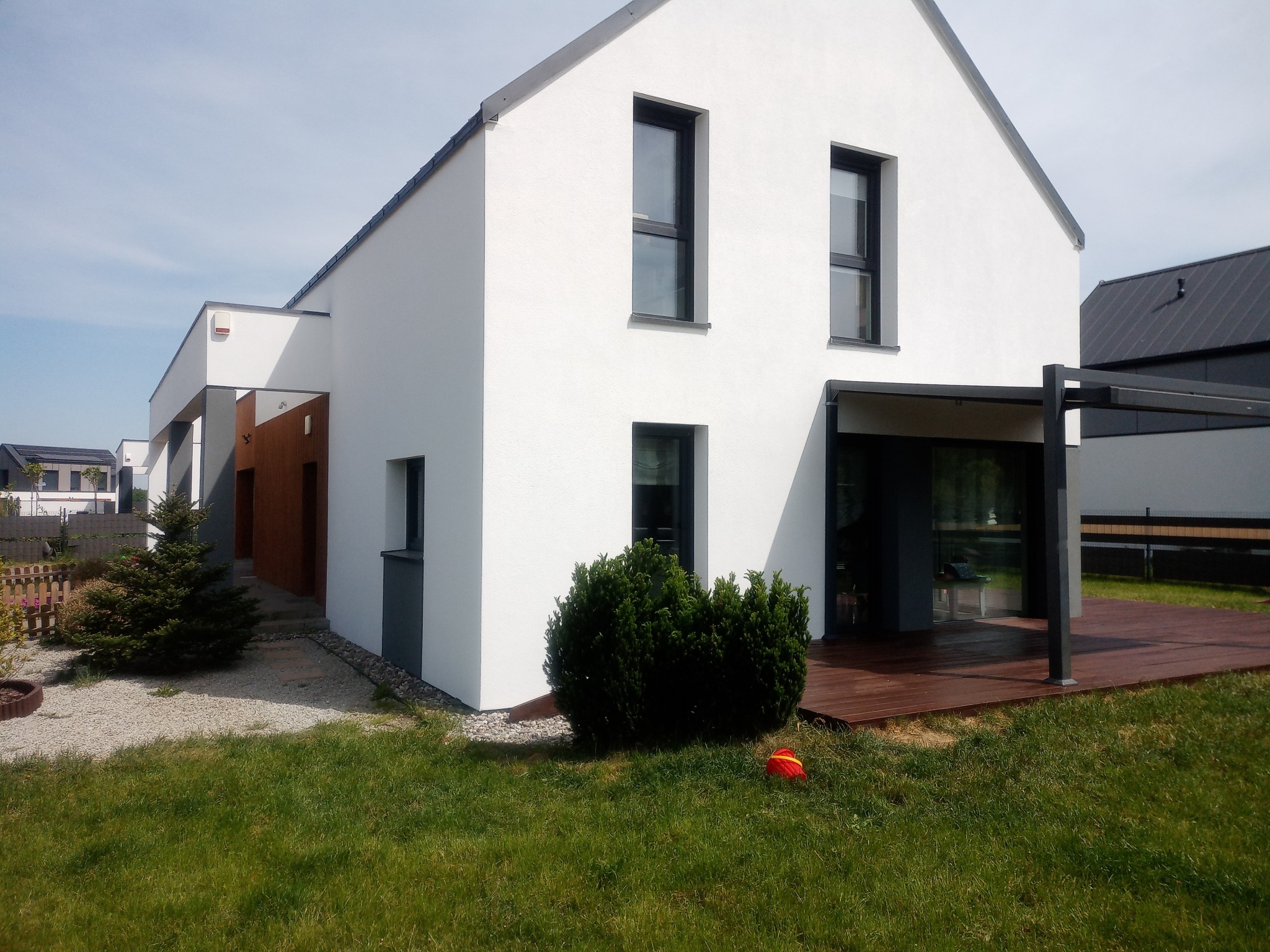 Nowoczesny, biały dom jednorodzinny z ciemnymi ramami okiennymi i drewnianym tarasem, widoczny z perspektywy ogrodu z niską zielenią i małą choinką.