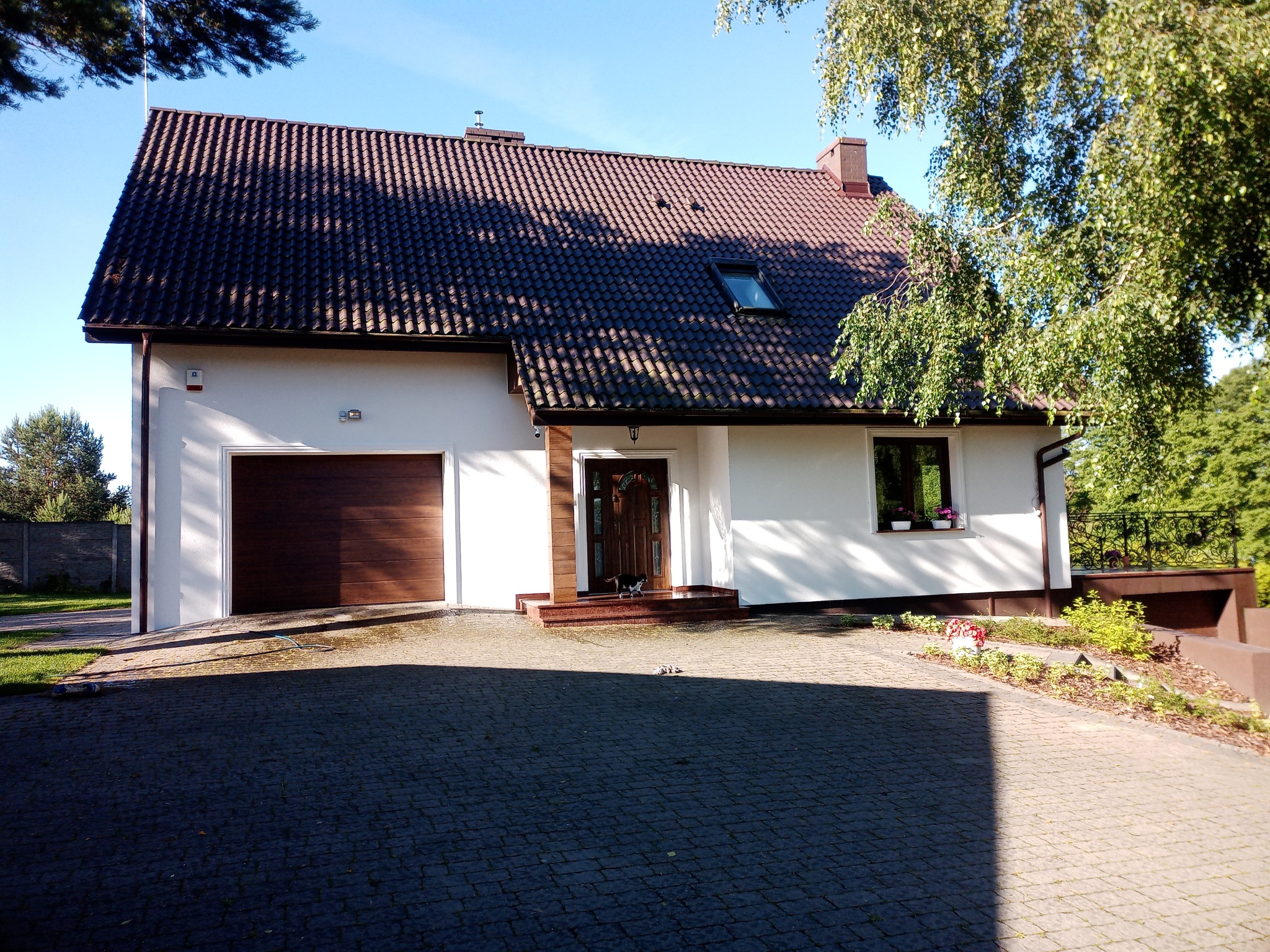 Biały dom jednorodzinny z brązową dachówką i bramą garażową, widok z brukowanego podjazdu w słoneczny dzień.