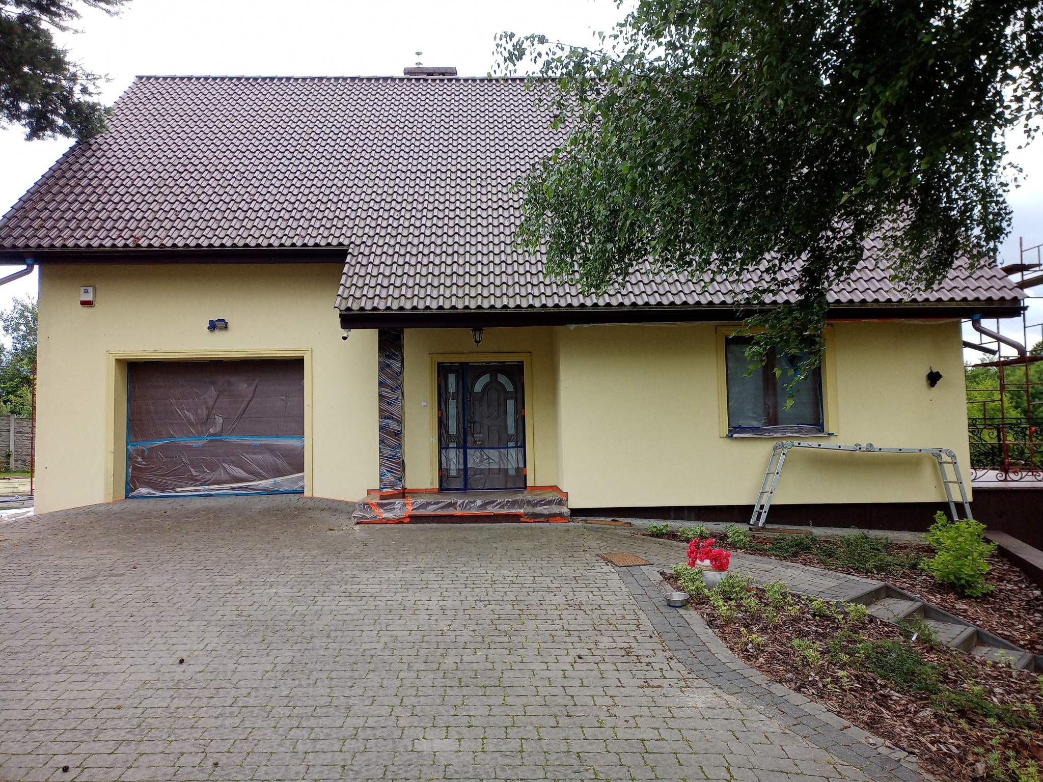 Żółty dom jednorodzinny z brązową dachówką w trakcie zabezpieczania elewacji przed pracami, okna i brama garażowa zakryte folią ochronną, widoczna drabina i rusztowanie w tle.