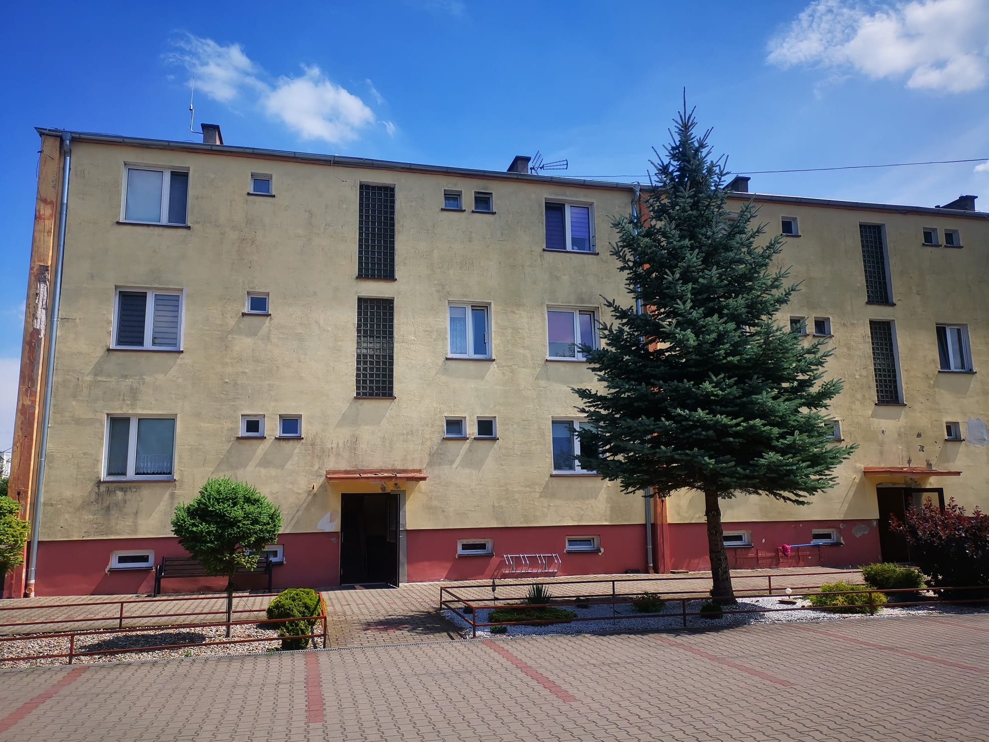 Żółta elewacja wielopiętrowego budynku z widocznymi śladami zabrudzeń i odprysków, z czerwonym pasem na dole, na tle błękitnego nieba z chmurami, przed budynkiem rosną drzewa.