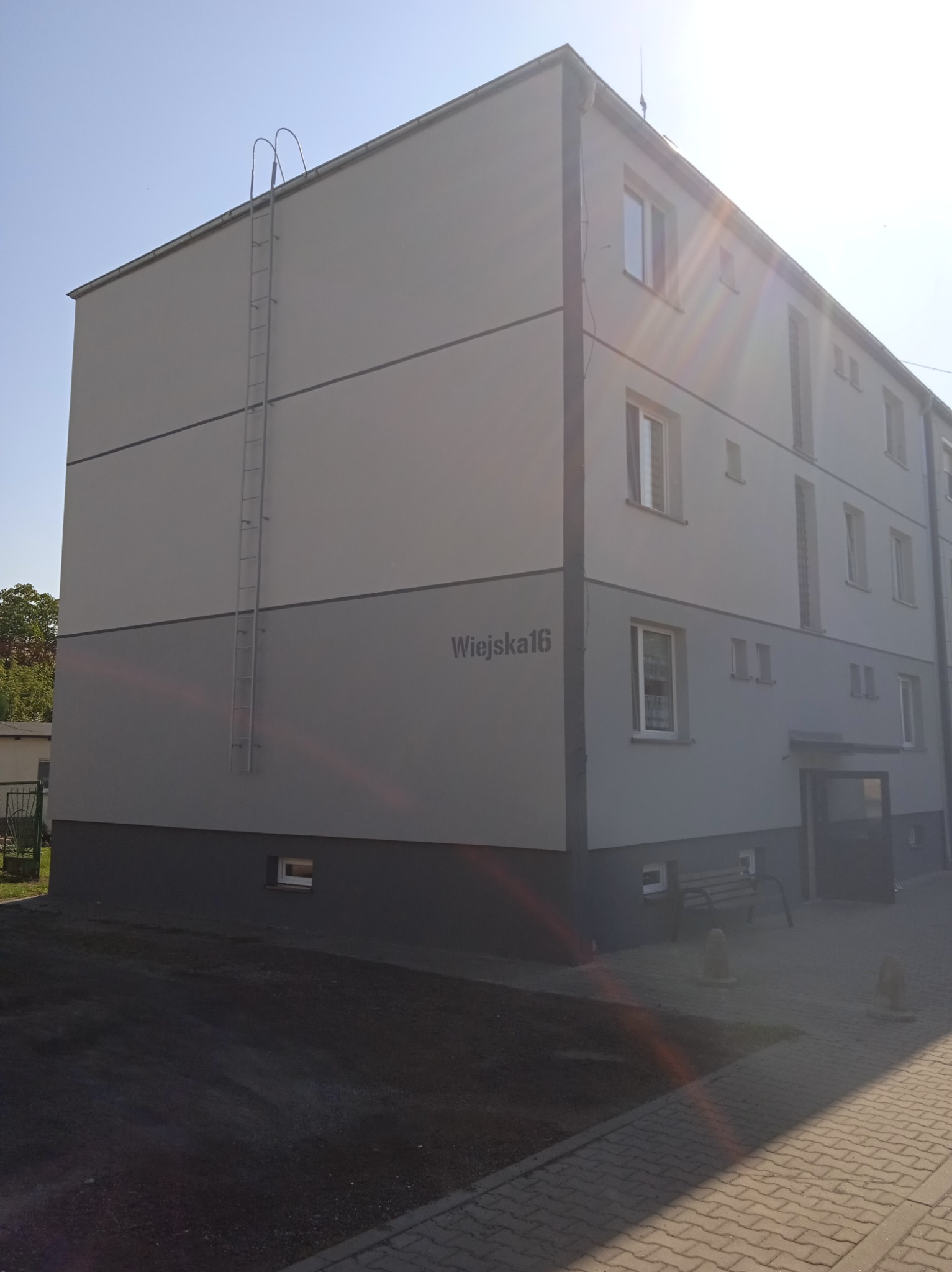 Odnowiony, trzypiętrowy budynek mieszkalny w Aleksandrowie Kujawskim z jasnoszarą elewacją, ciemnoszarym cokołem i numerem 'Wiejska 16', wyposażony w zewnętrzną drabinę przeciwpożarową.