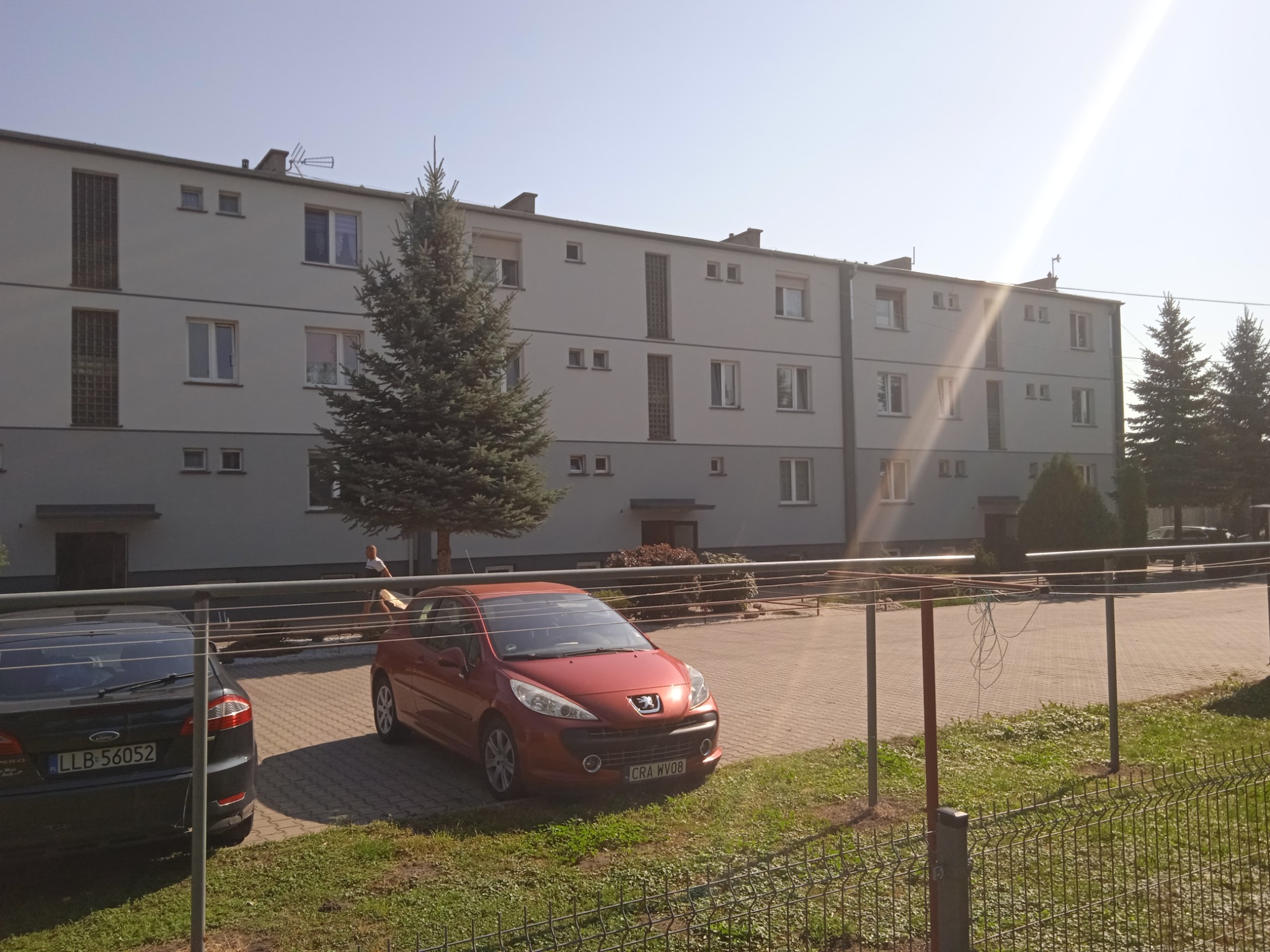 Trzypiętrowy budynek mieszkalny z szarą elewacją, widoczny zza ogrodzenia, z zaparkowanymi samochodami i suszarką na pranie na pierwszym planie.