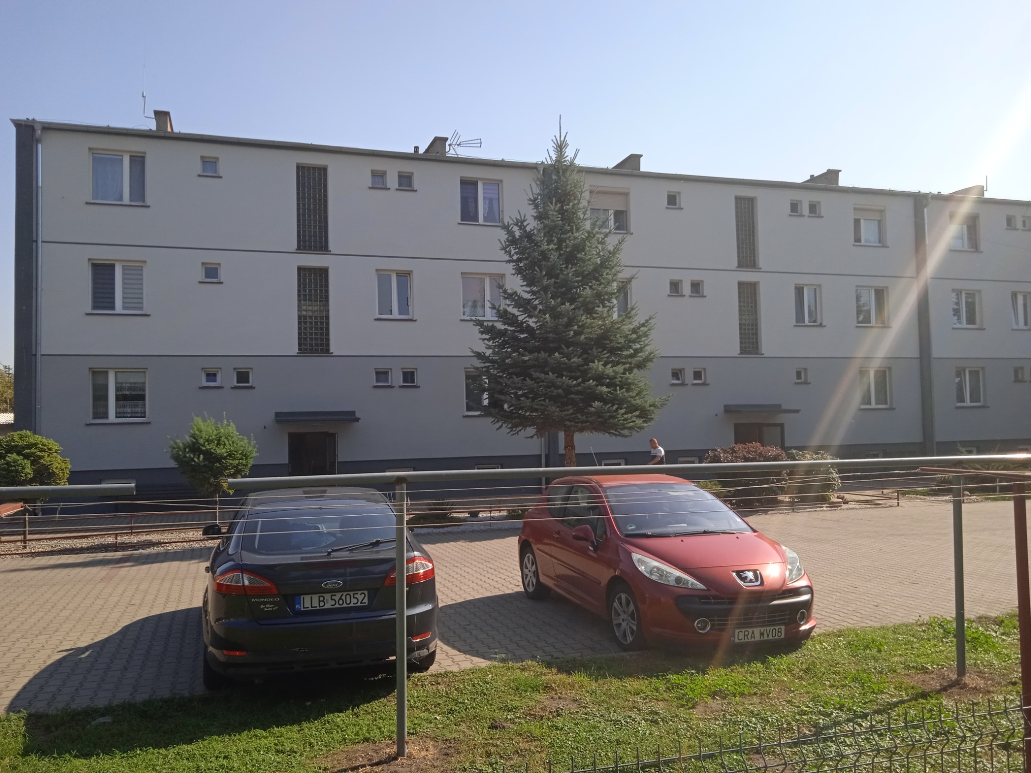 Trzypiętrowy budynek mieszkalny z szarą elewacją, widoczny z poziomu parkingu z dwoma samochodami, przed budynkiem rośnie duża choinka.