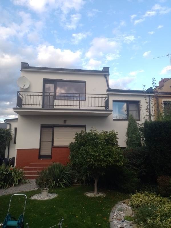 Dwupiętrowy dom z jasną elewacją, balkonem z czarną balustradą i czerwoną ceglaną podstawą, widok z ogrodu z zielonym trawnikiem i drzewami.