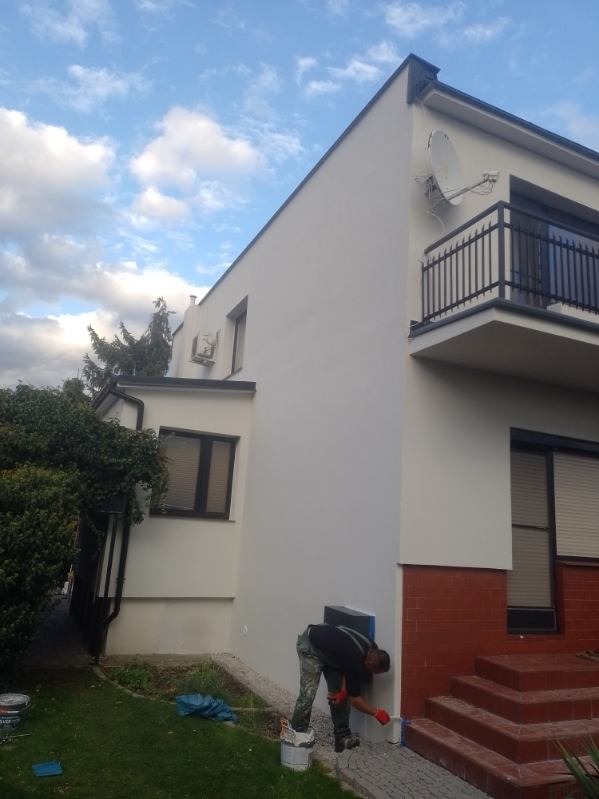 Mężczyzna w kamuflażowych spodniach maluje dolną część elewacji domu na biało, używając pędzla i stojąc obok wiadra z farbą, z widocznymi elementami czerwonej cegły i schodami.