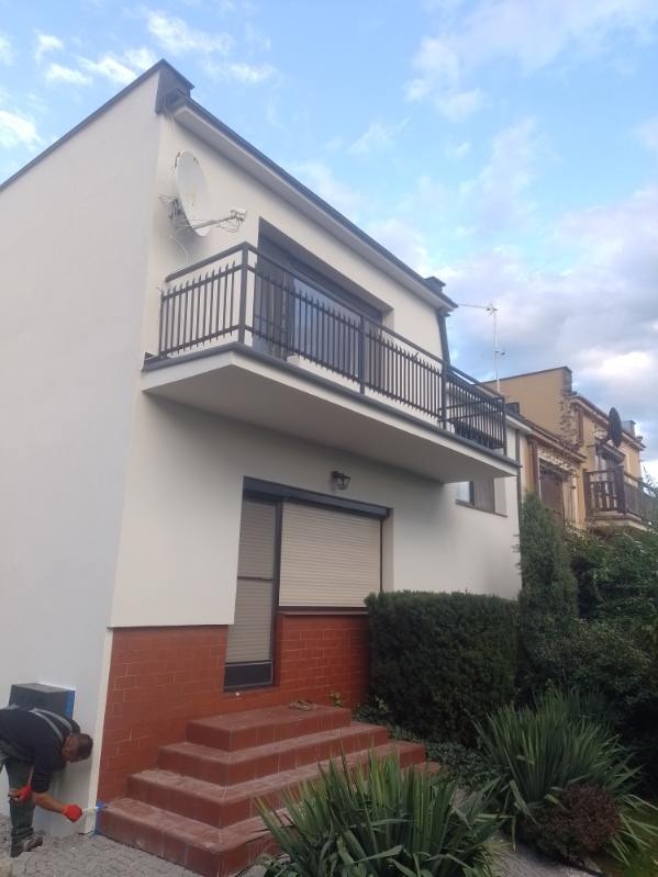 Pracownik maluje dolną część elewacji domu z ceglaną podmurówką, widoczny balkon z czarną balustradą i satelita na ścianie.