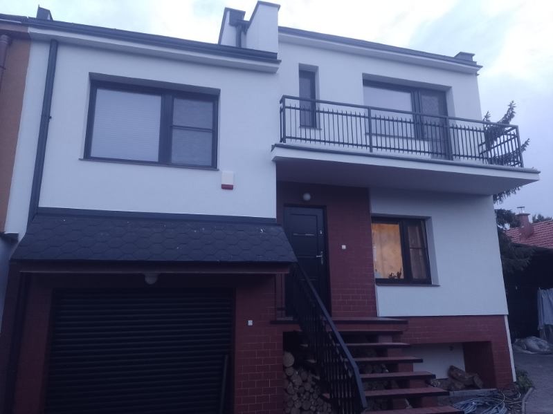 Dwukondygnacyjny dom z balkonem i garażem po renowacji elewacji, z widocznym fragmentem muru z cegły klinkierowej u podstawy i stosem drewna na opał obok schodów wejściowych.