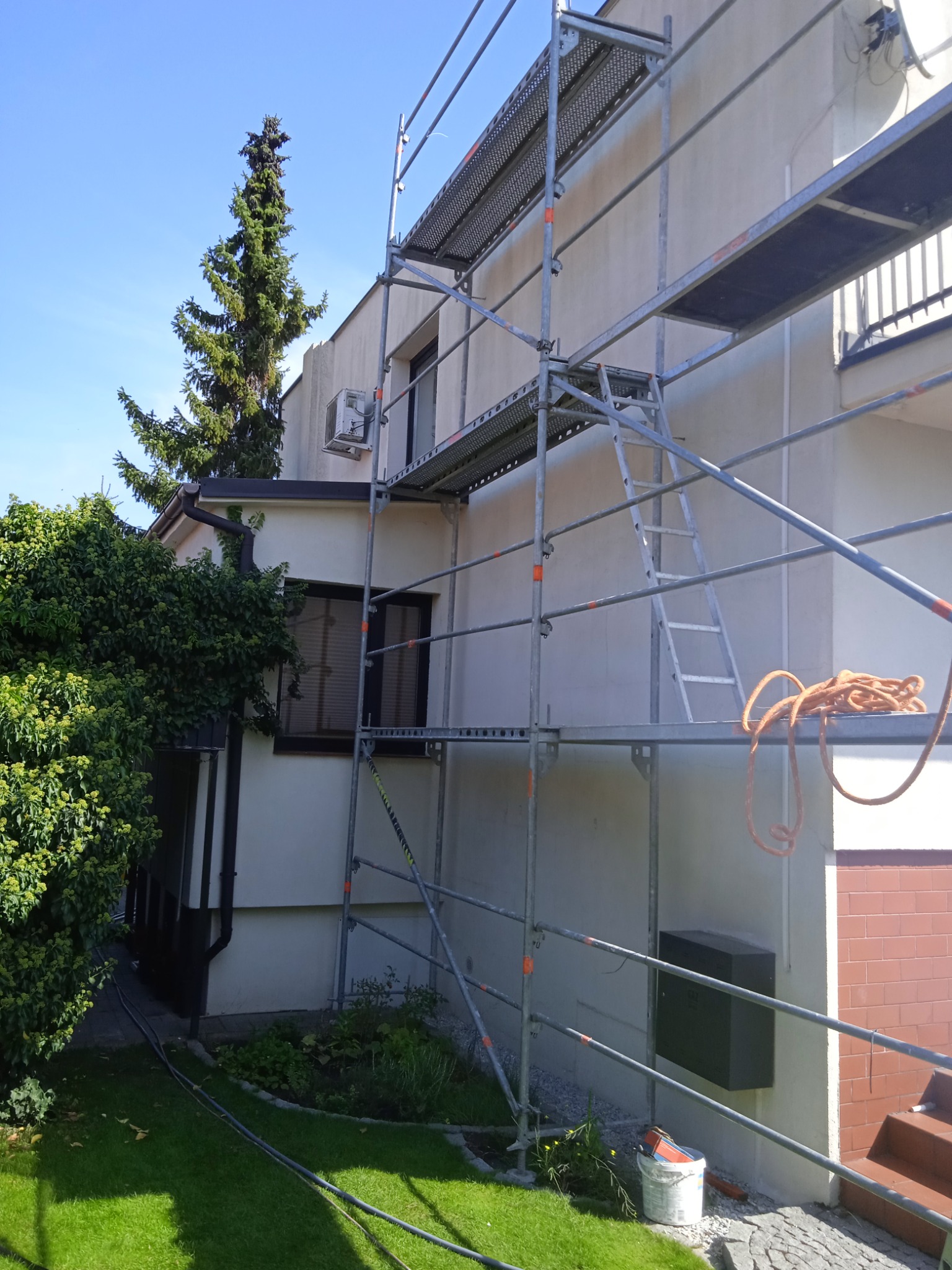 Elewacja domu w trakcie prac: widoczne rusztowanie, fragment ocieplonej ściany, okno z klimatyzatorem i zwisająca lina.