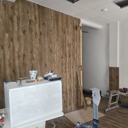 L.W.Hall Łukasz Wójcik - Montaż paneli ściennych imitujących drewno, widoczny fragment ściany z zamontowanymi panelami, biała szafka z narzędziami i materiałami budowlanymi, oraz drabina w tle, sugerująca trwające prace...