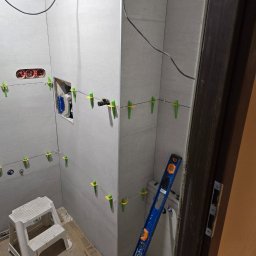 L.W.Hall Łukasz Wójcik - Łazienka w trakcie remontu: szare płytki ścienne, poziomica oparta o ścianę, plastikowy stołek, widoczne gniazdka elektryczne i otwór na instalację wodną zabezpieczony folią, system poziomowania...