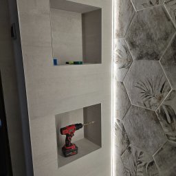 L.W.Hall Łukasz Wójcik - Wnęka w szarej ścianie wykończonej płytkami z motywem liści, z wiertarką akumulatorową w środku, gotowa do wiercenia.