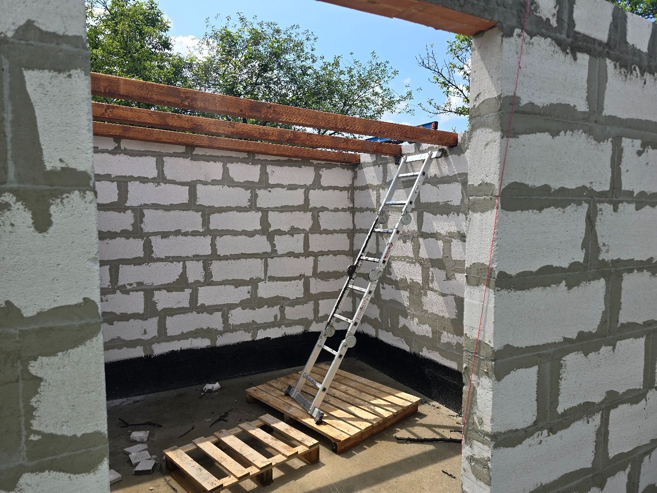 Wznoszenie ścian z bloczków betonowych Ytong, widoczne drewniane belki stropowe i drabina oparta o ścianę na paletach. Izolacja przeciwwilgociowa przy podłożu.
