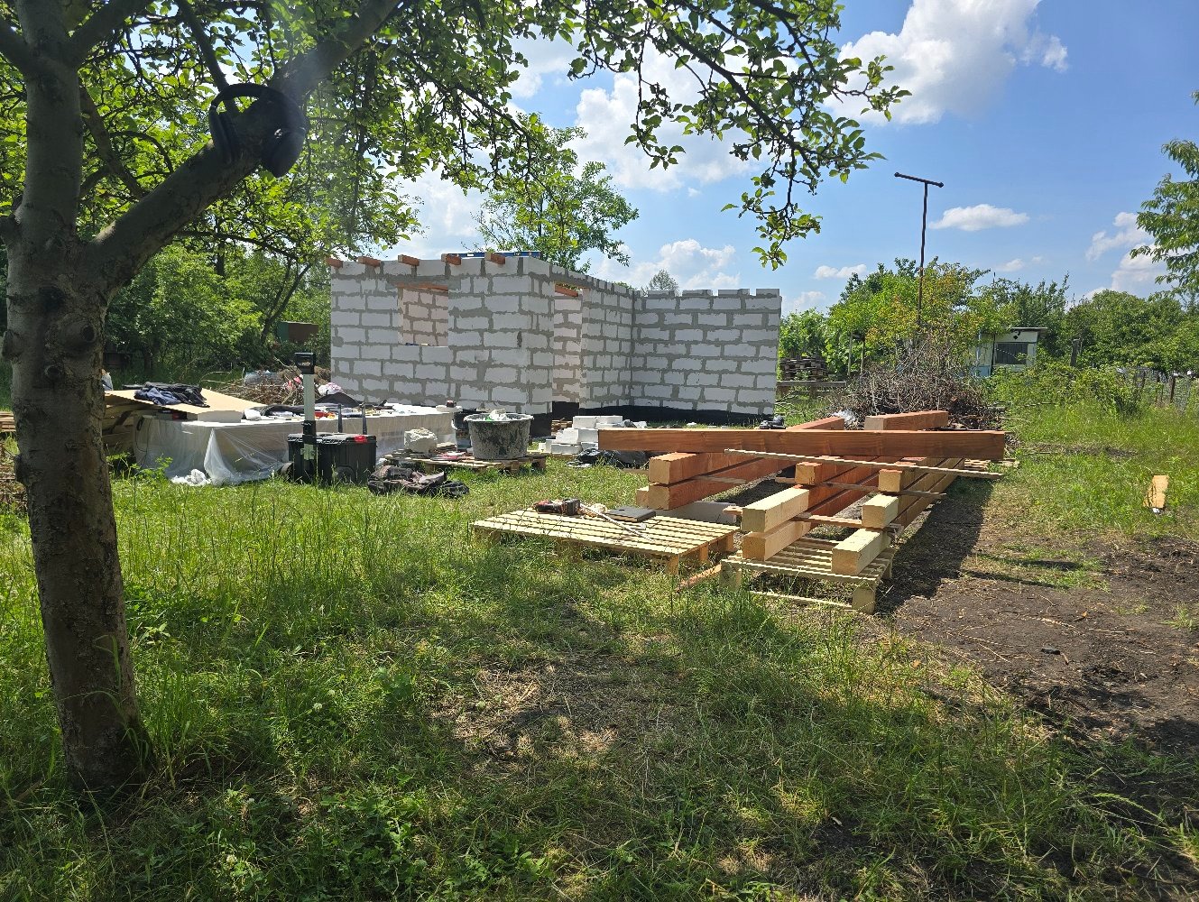 Wznoszenie ścian z bloczków betonowych na fundamencie w otoczeniu zieleni, widoczne drewniane belki i palety przygotowane do dalszych prac budowlanych.