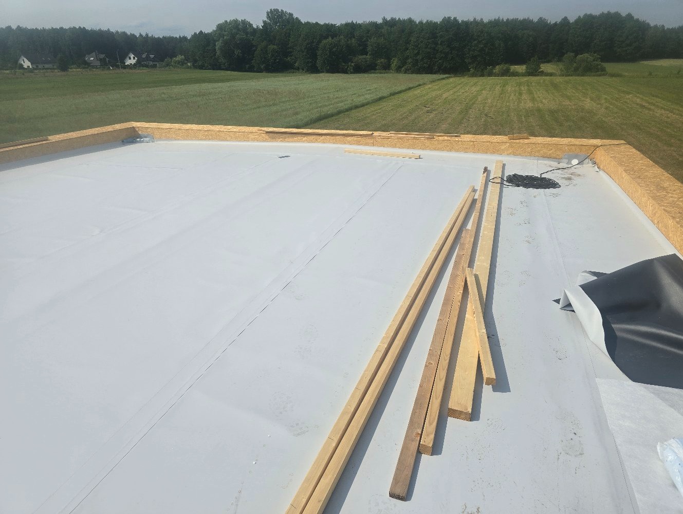 Biała membrana dachowa na płaskim dachu, z drewnianą obróbką krawędzi i przygotowanymi łatami, widok na zielone pole i las w tle.