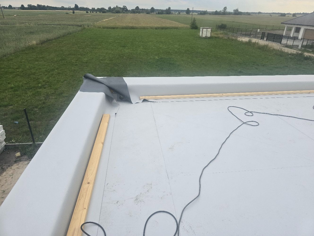 Widok z góry na płaski dach pokryty szarą membraną, z widocznymi drewnianymi listwami, luźno zwisającym czarnym kablem i fragmentem niezabezpieczonej krawędzi dachu z wystającą czarną folią, na tle...