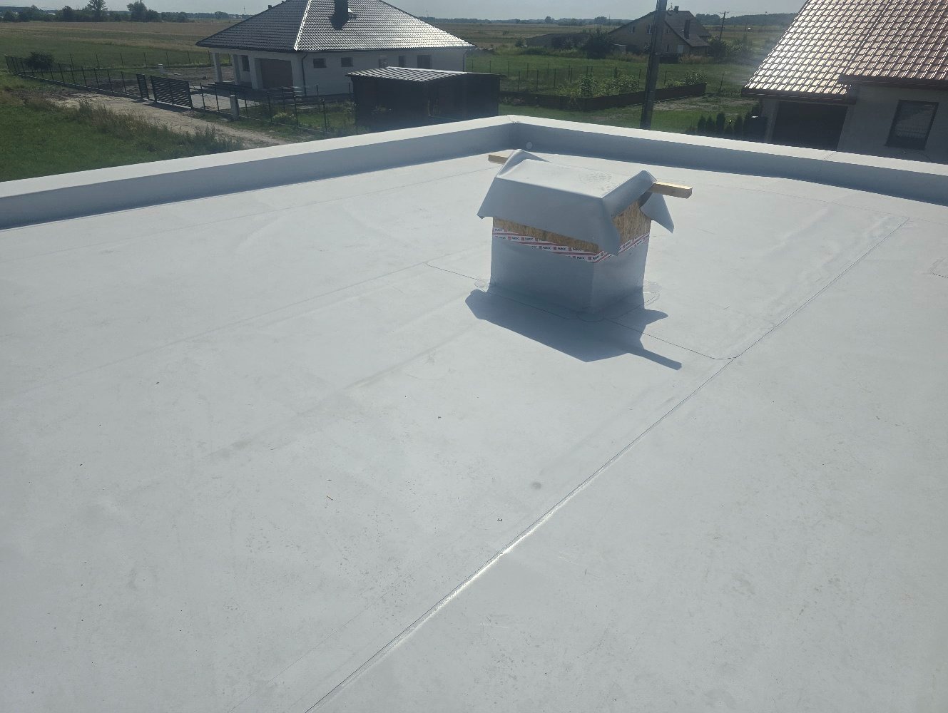Szary płaski dach z widocznym kominem zabezpieczonym folią i taśmą, w tle domy jednorodzinne.