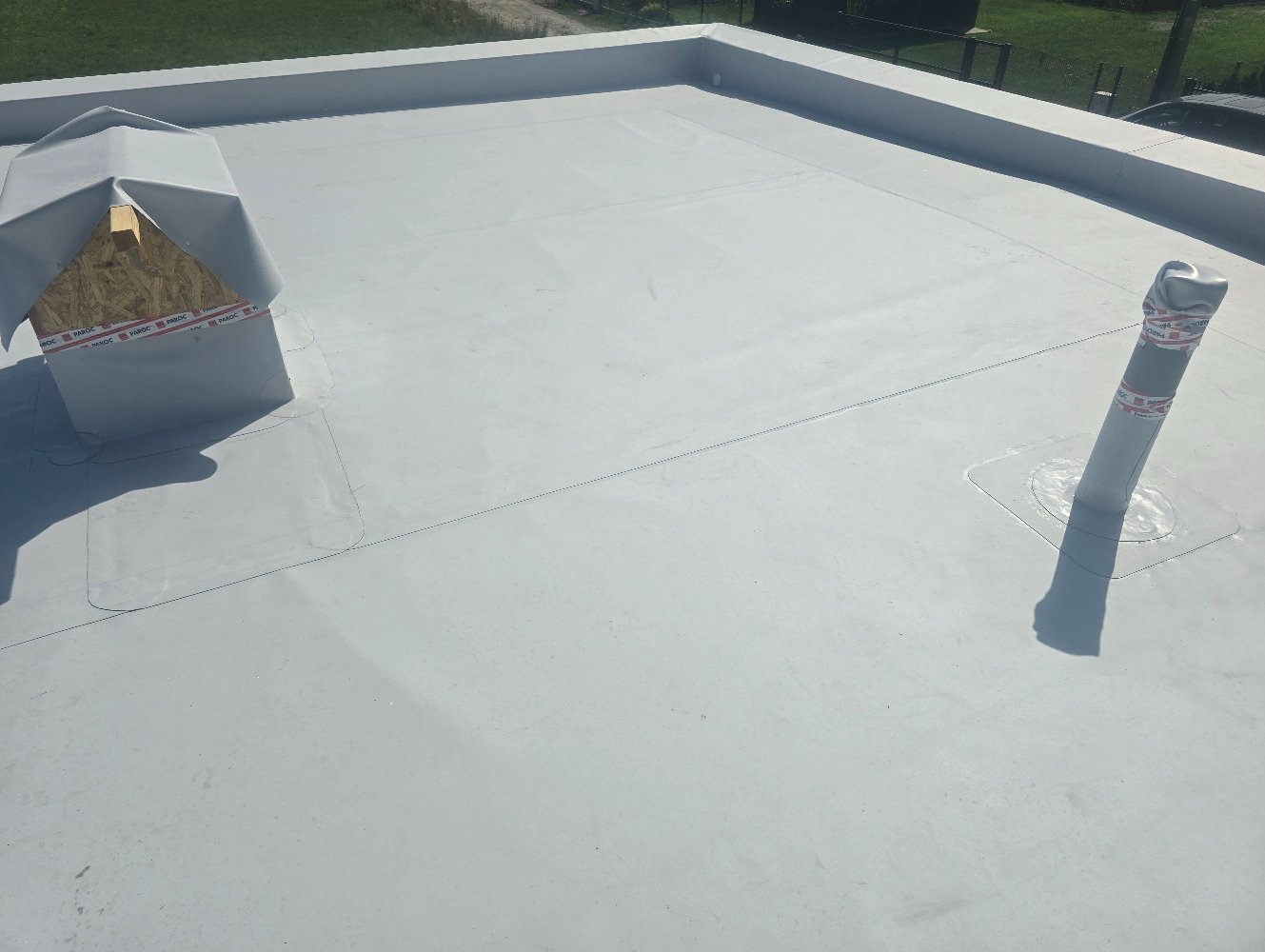 Nowo położona, jasnoszara membrana dachowa na płaskim dachu z widocznymi detalami, takimi jak kominek wentylacyjny zabezpieczony taśmą i rura odpowietrzająca.