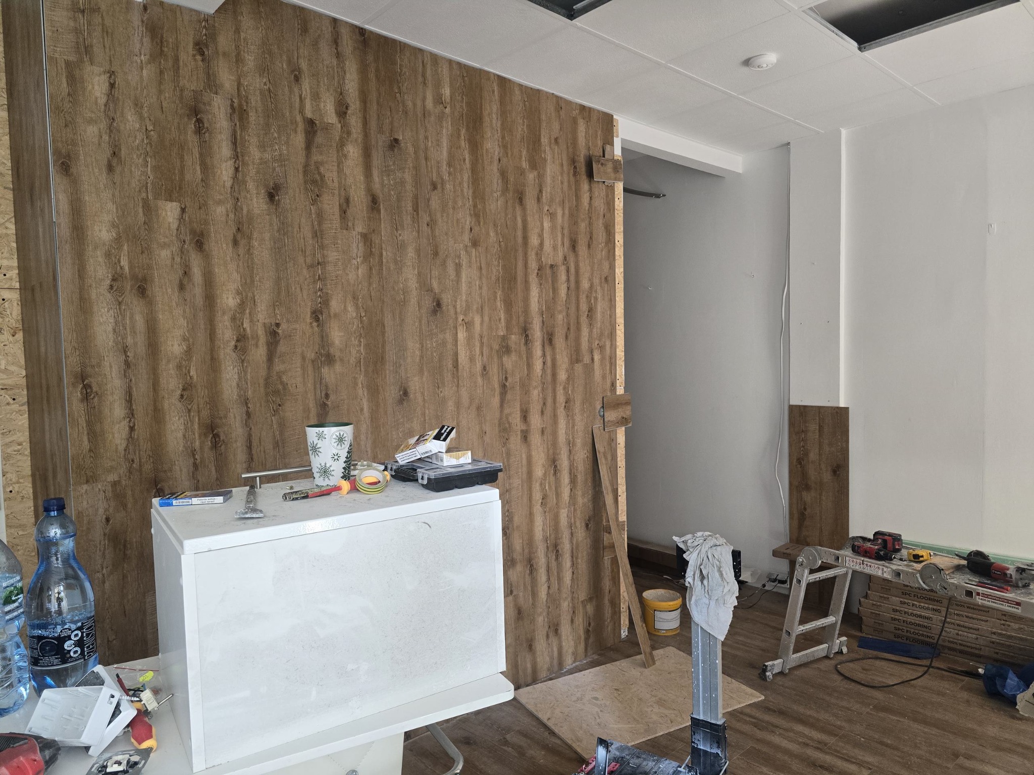 Montaż paneli ściennych imitujących drewno, widoczny fragment ściany z zamontowanymi panelami, biała szafka z narzędziami i materiałami budowlanymi, oraz drabina w tle, sugerująca trwające prace...