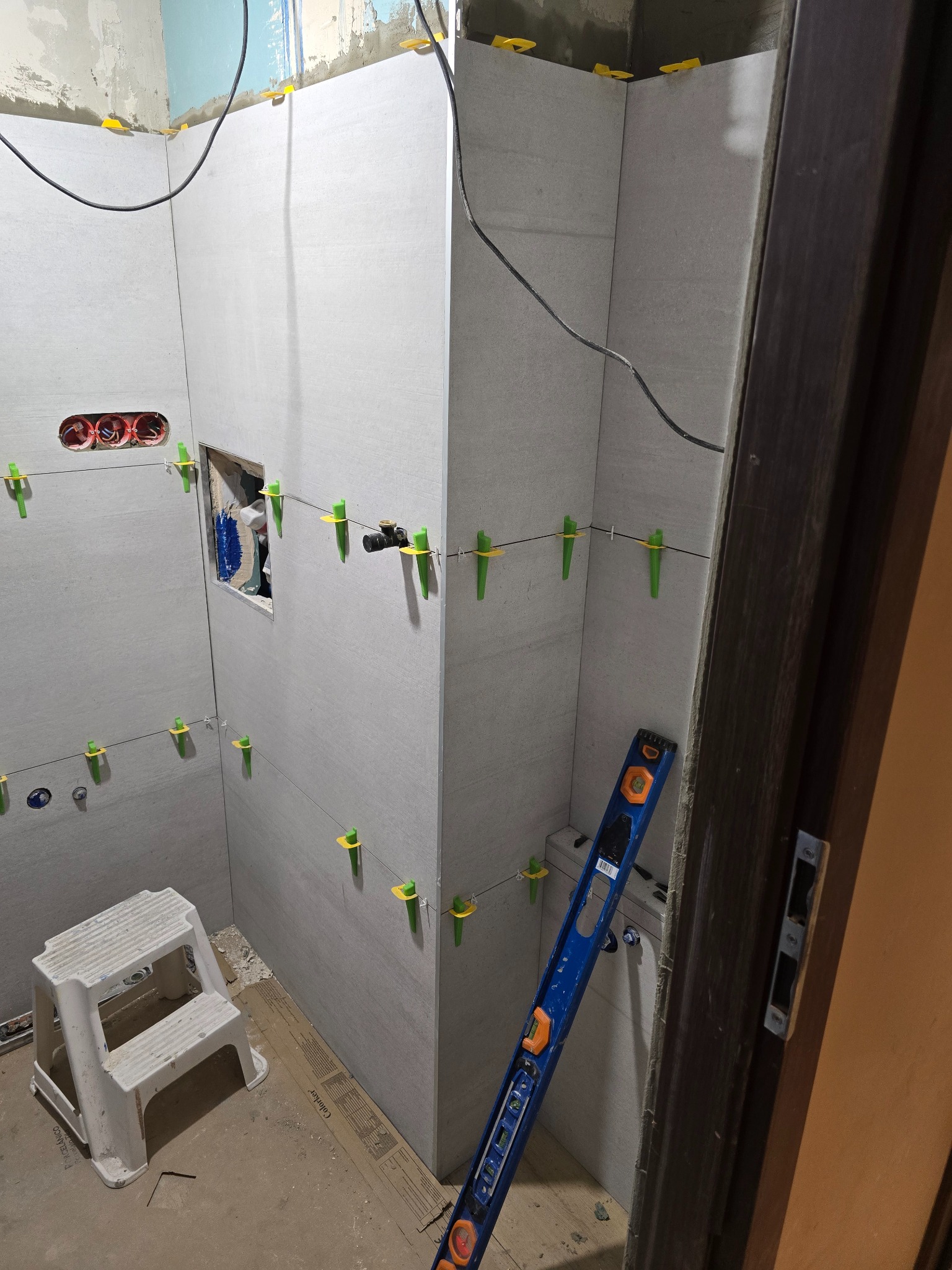 Łazienka w trakcie remontu: szare płytki ścienne, poziomica oparta o ścianę, plastikowy stołek, widoczne gniazdka elektryczne i otwór na instalację wodną zabezpieczony folią, system poziomowania...