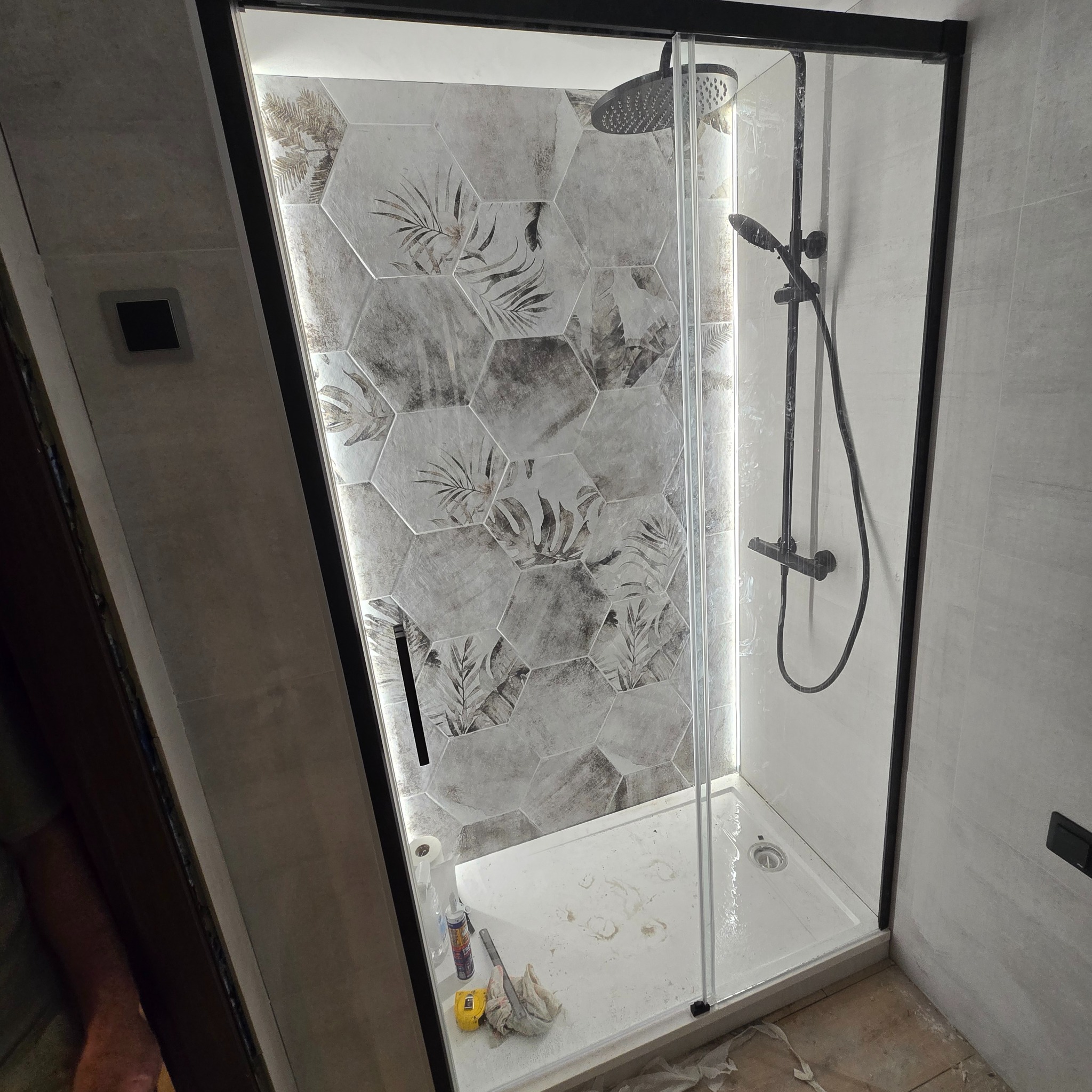 Nowoczesna kabina prysznicowa z czarną ramą i deszczownicą, heksagonalne płytki z motywem liści, podświetlenie LED i widoczne narzędzia podczas montażu.