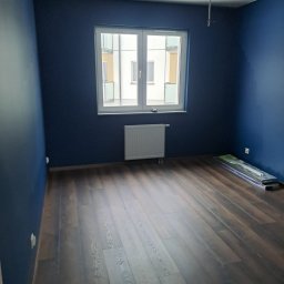 Kar-Bud - Pokój po malowaniu na granatowo z drewnianą podłogą. Widoczne okno z grzejnikiem i listwy przypodłogowe w opakowaniu. Kabel zwisa z sufitu.