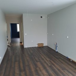 Kar-Bud - Wnętrze po remoncie: ciemne panele podłogowe, szare ściany z widocznymi instalacjami. W tle widoczny fragment innego pomieszczenia z ciemną ścianą.