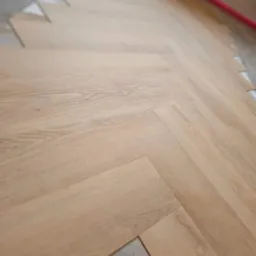 Układanie jasnego parkietu w jodełkę francuską, widoczne docięte elementy i podłoże pod parkiet.