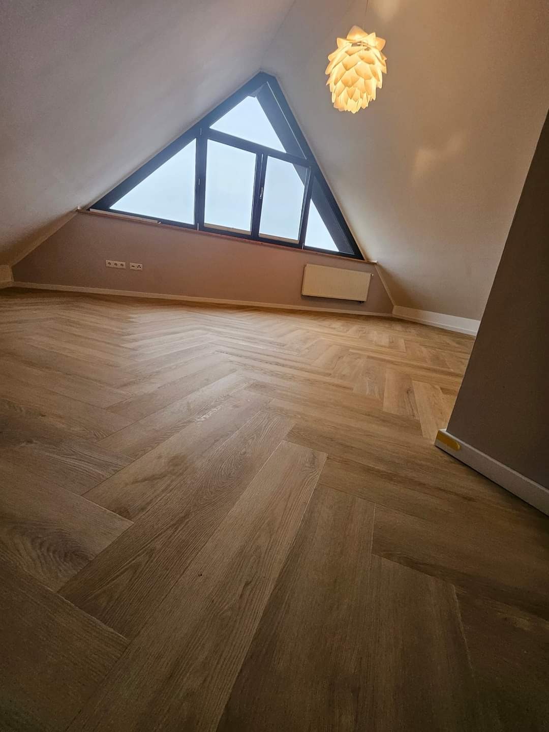 Jasny parkiet ułożony w jodełkę w pokoju na poddaszu z trójkątnym oknem i designerską lampą.