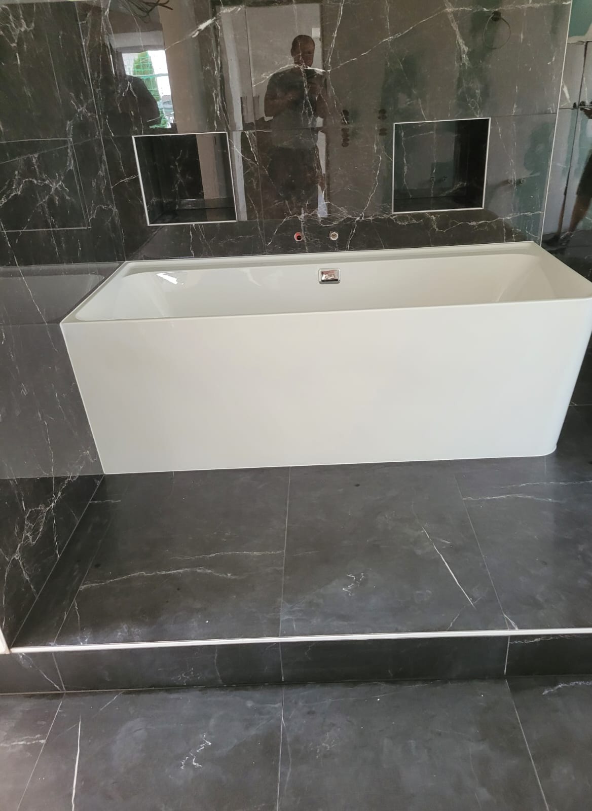Nowoczesna, prostokątna wanna w łazience wyłożonej czarnym marmurem z białymi żyłami; w tle widać lustro i wnęki w ścianie.