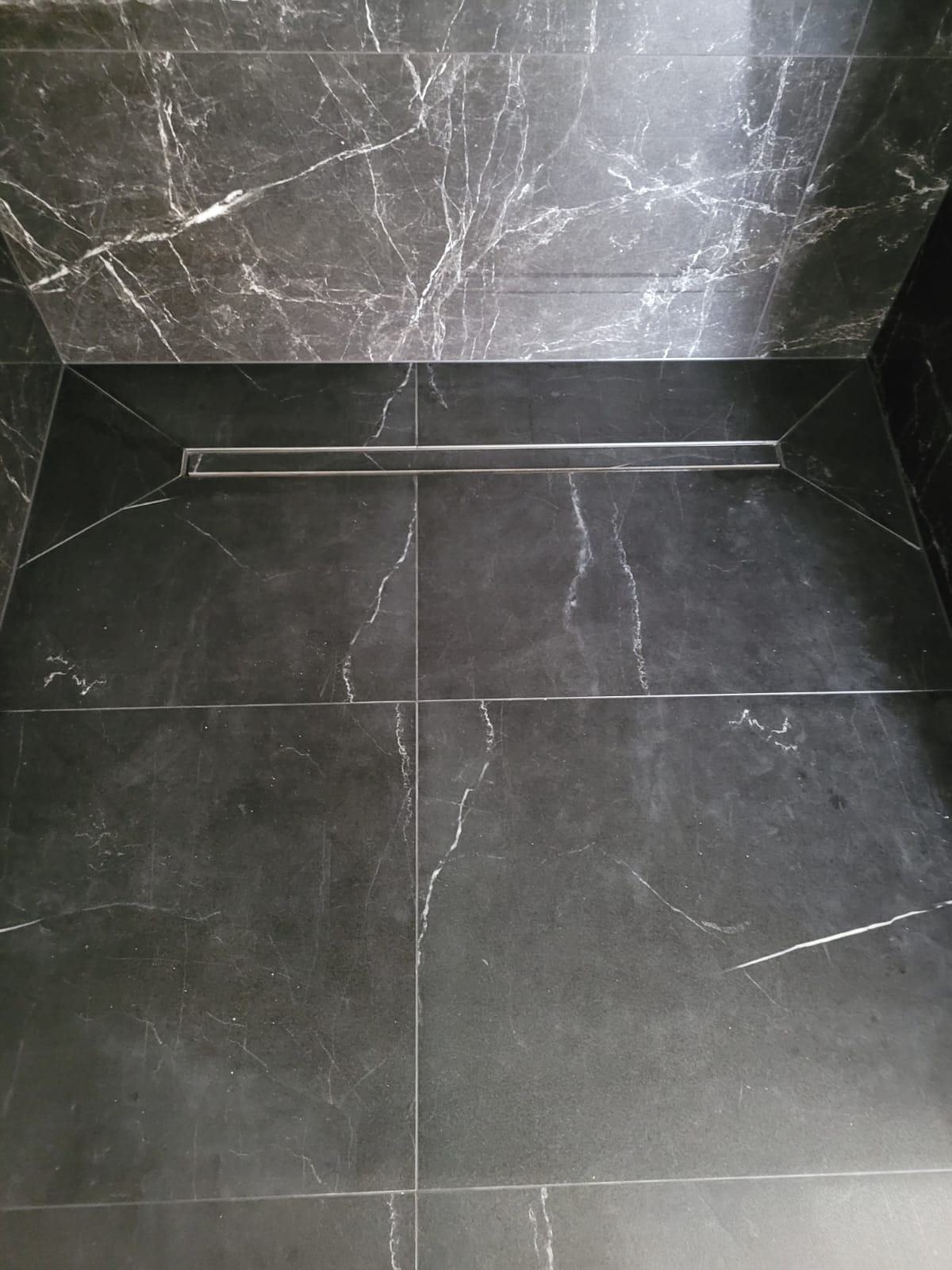 Wykończona kabina prysznicowa z czarnymi płytkami imitującymi marmur i liniowym odpływem podłogowym ze stali nierdzewnej.