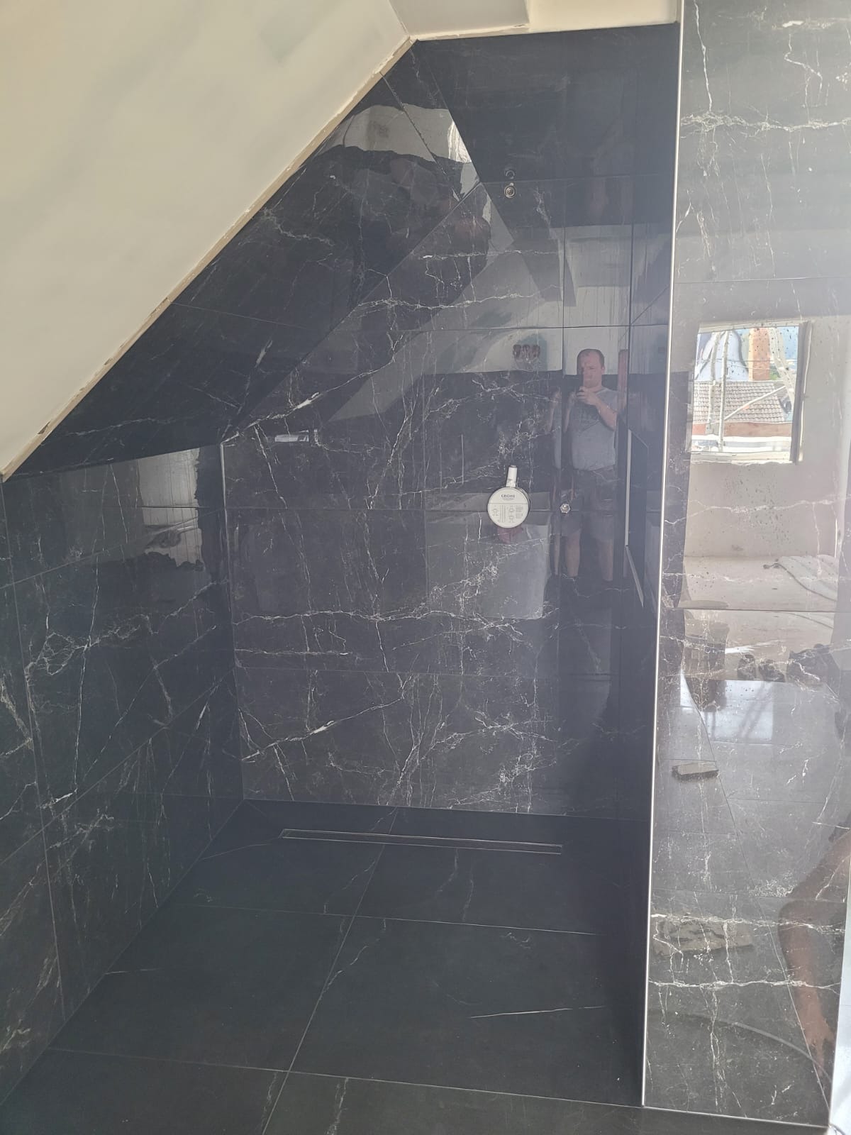 Wykończona kabina prysznicowa w łazience na poddaszu, wyłożona czarnymi płytkami imitującymi marmur z widocznym odpływem liniowym i zamontowaną baterią Grohe; w lustrzanej powierzchni odbija się...