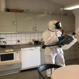 Osoba w kombinezonie ochronnym przeprowadza zamgławianie w kuchni, używając profesjonalnego urządzenia do rozpylania środka dezynfekującego.