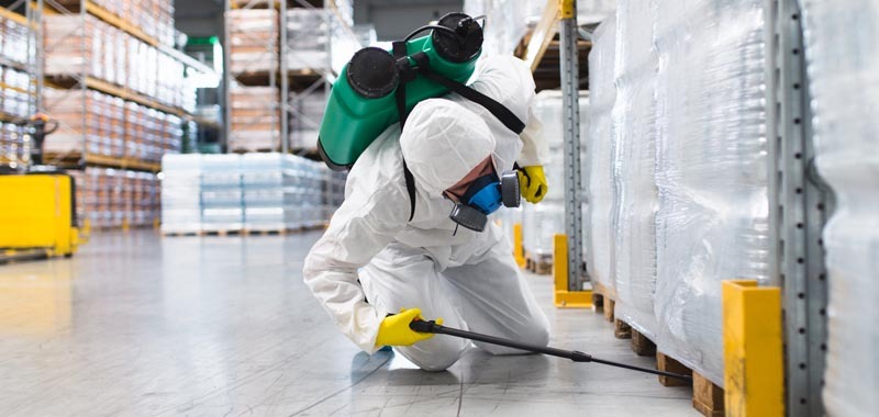 Pracownik w kombinezonie ochronnym i żółtych rękawicach, z maską na twarzy i opryskiwaczem na plecach, rozpyla preparat w magazynie, widoczne regały z zapakowanymi towarami.