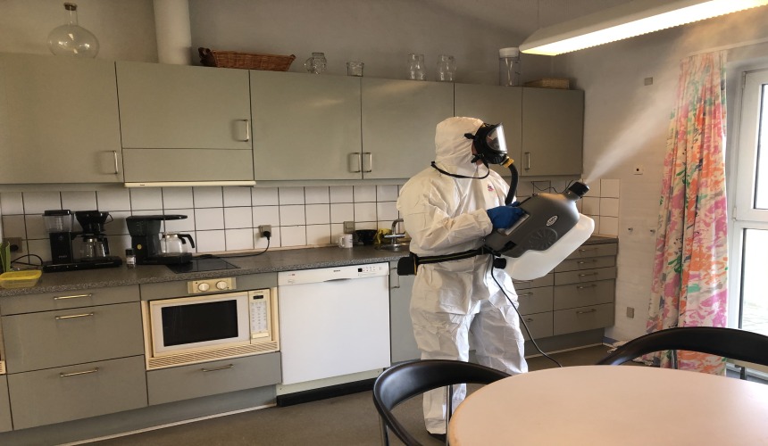 Osoba w kombinezonie ochronnym przeprowadza zamgławianie w kuchni, używając profesjonalnego urządzenia do rozpylania środka dezynfekującego.