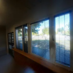 Trzy okna z poziomymi żaluzjami aluminiowymi, widok na zewnątrz z drzewami i placem zabaw w słoneczny dzień.