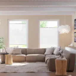 Jasne wnętrze salonu z dwoma oknami przesłoniętymi białymi plisami, beżowa sofa narożna, drewniane stoliki pomocnicze i puszysty dywan.