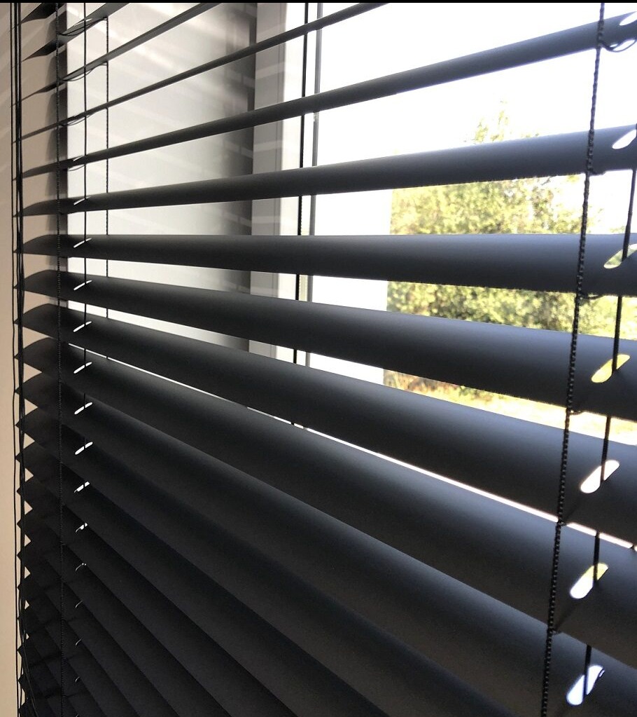 Częściowo zamknięte, matowe, ciemne żaluzje aluminiowe w oknie, widok na zieleń na zewnątrz.