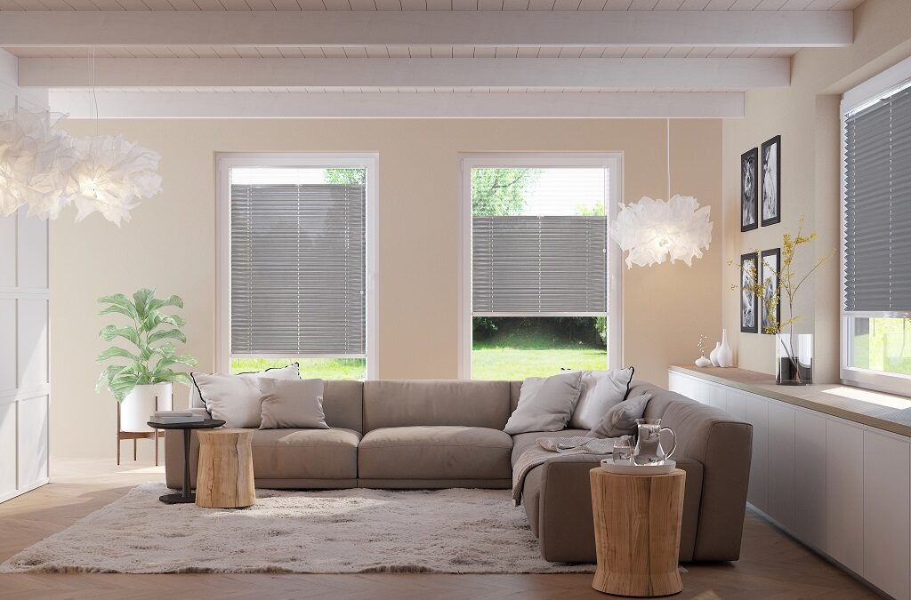 Nowoczesny salon z beżową sofą narożną i szarymi plisowanymi roletami okiennymi, widok na zieleń za oknem, dekoracyjne lampy i drewniane stoliki kawowe.