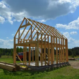 Wznoszenie dwukondygnacyjnej konstrukcji szkieletowej domu z drewna na betonowym fundamencie, z widocznymi belkami stropowymi i dachowymi, na tle zielonej trawy i błękitnego nieba z chmurami.