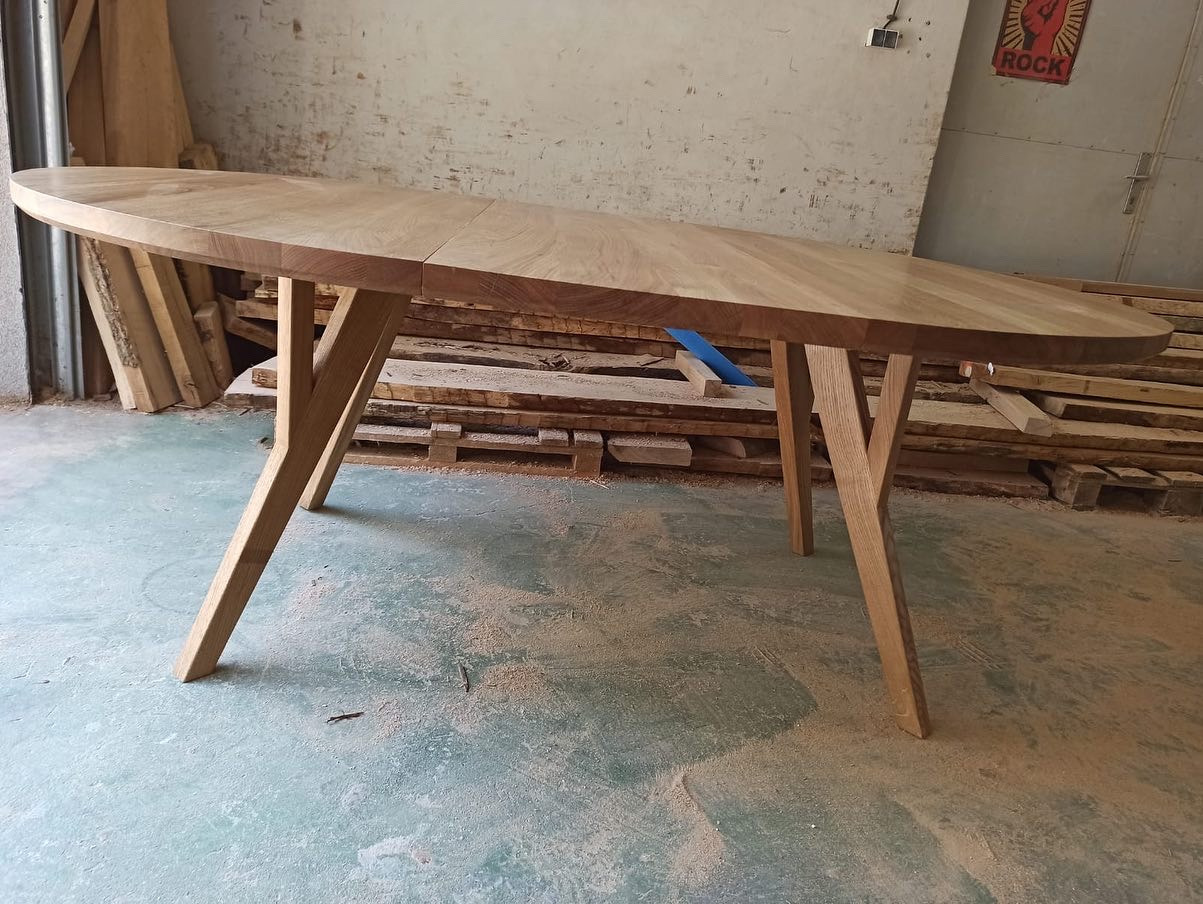 Rozkładany, owalny stół z jasnego drewna na skośnych nogach, widoczny w warsztacie stolarskim, tło stanowią deski i plakat z motywem rockowym.