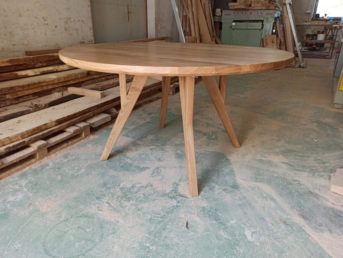 Okrągły stół z jasnego drewna z trzema nogami, stojący w warsztacie stolarskim na podłodze pokrytej pyłem drzewnym, z widocznymi deskami i maszynami w tle.