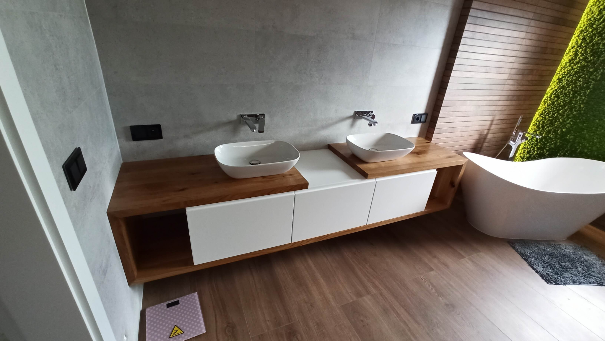 Nowoczesna łazienka z drewnianym blatem pod dwie umywalki nablatowe, białe szafki, wolnostojąca wanna i ściana z mchu dekoracyjnego.