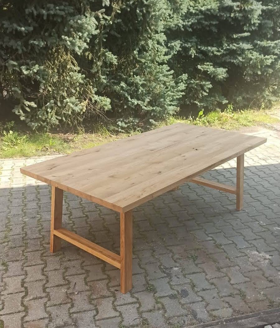 Prostokątny stół z jasnego drewna, z widoczną strukturą słojów, ustawiony na kostce brukowej przed zielonym krzewem.