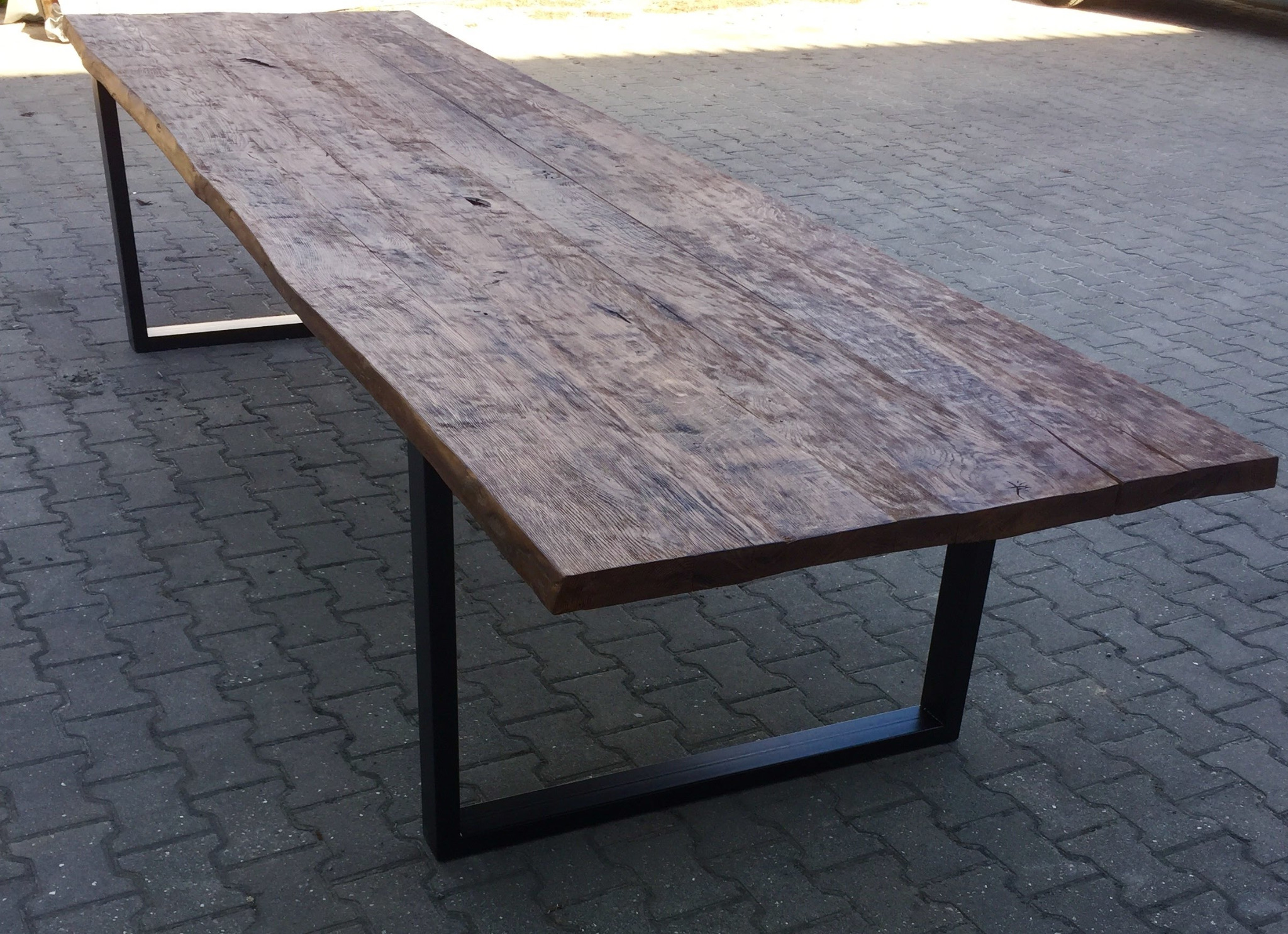 Długi, rustykalny blat stołu z ciemnego drewna o nieregularnych krawędziach, wsparty na czarnych, metalowych nogach w kształcie prostokątnych ram, ustawiony na szarej kostce brukowej.