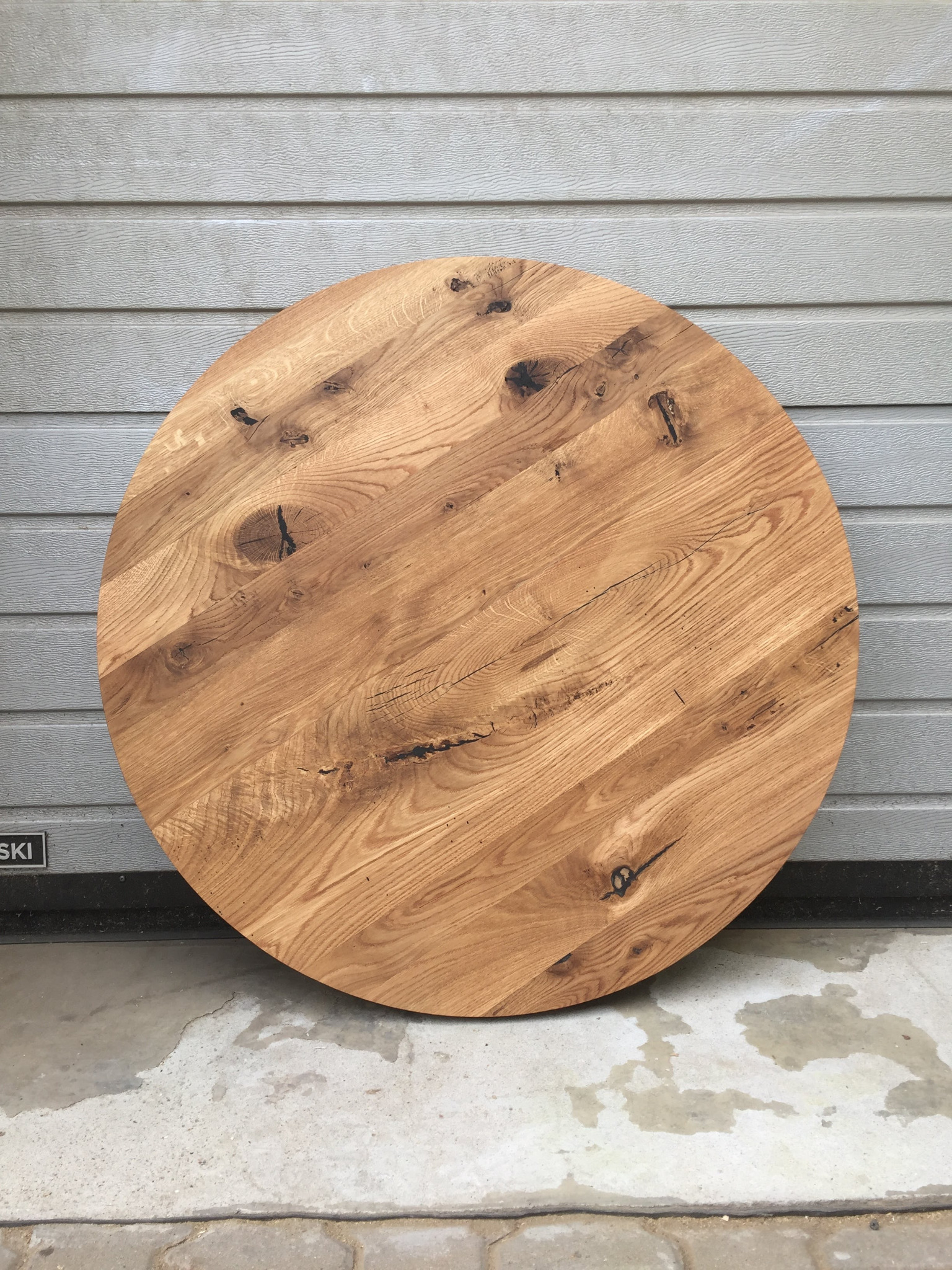 Okrągły blat drewniany z wyraźnym usłojeniem i naturalnymi sękami, oparty o szarą ścianę z paneli i betonową podłogę z plamami.