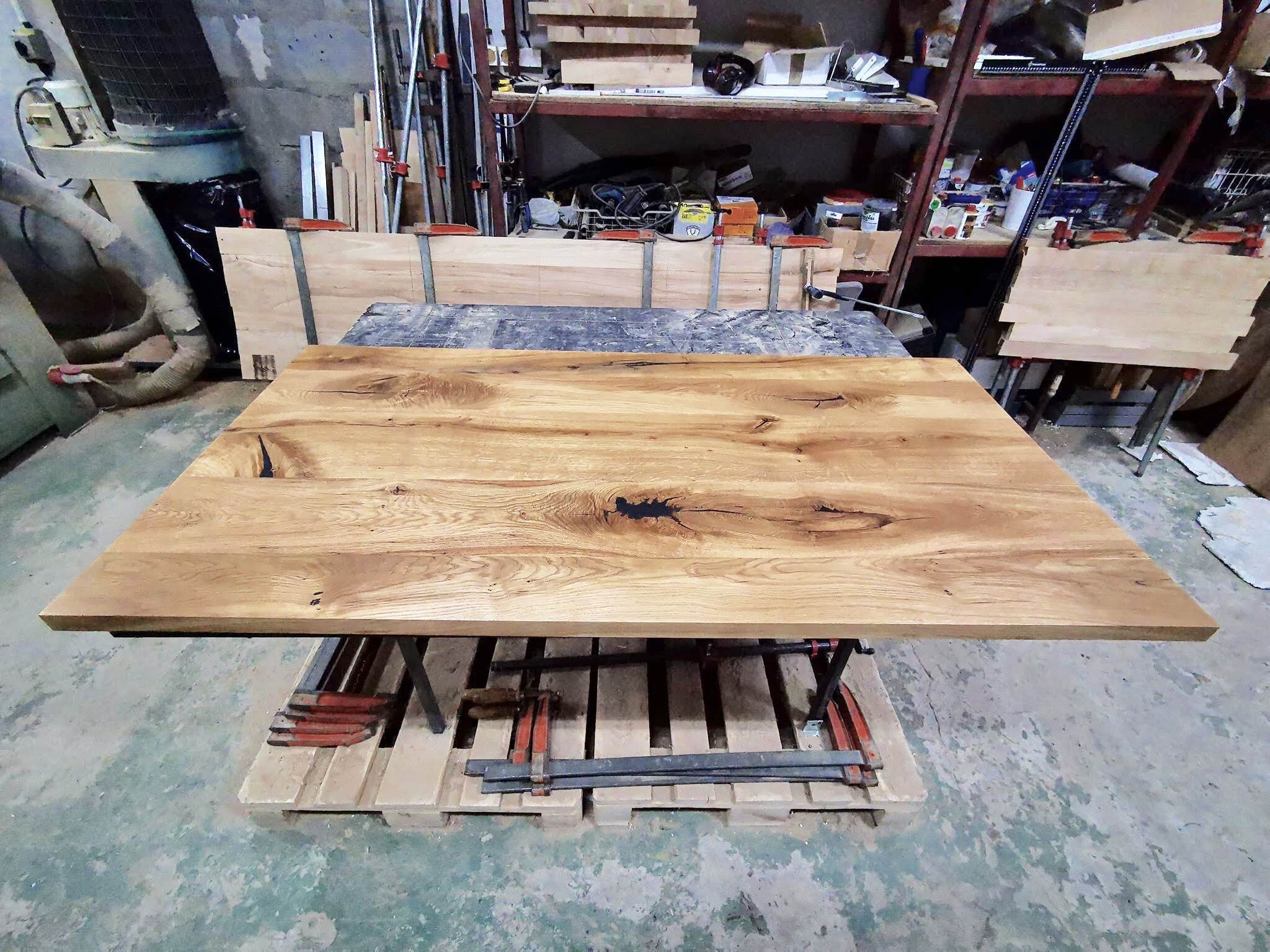 Surowy blat z jasnego drewna dębowego z widocznymi sękami i naturalnym usłojeniem, leżący na drewnianej palecie w warsztacie, otoczony narzędziami i sprzętem stolarskim.