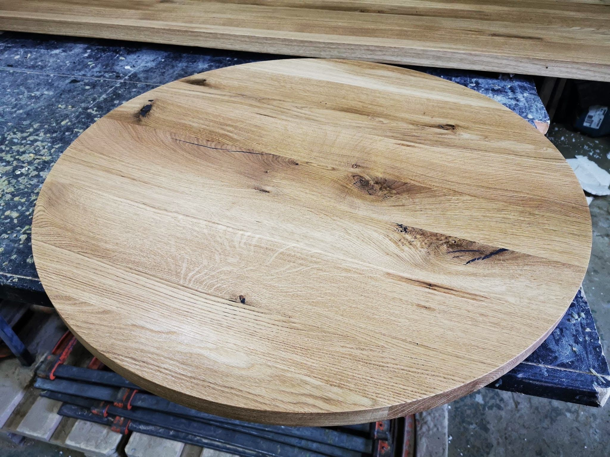 Okrągły blat z jasnego drewna dębowego z widocznymi sękami, leżący na roboczym stole w warsztacie, z klamrami stolarskimi w tle.
