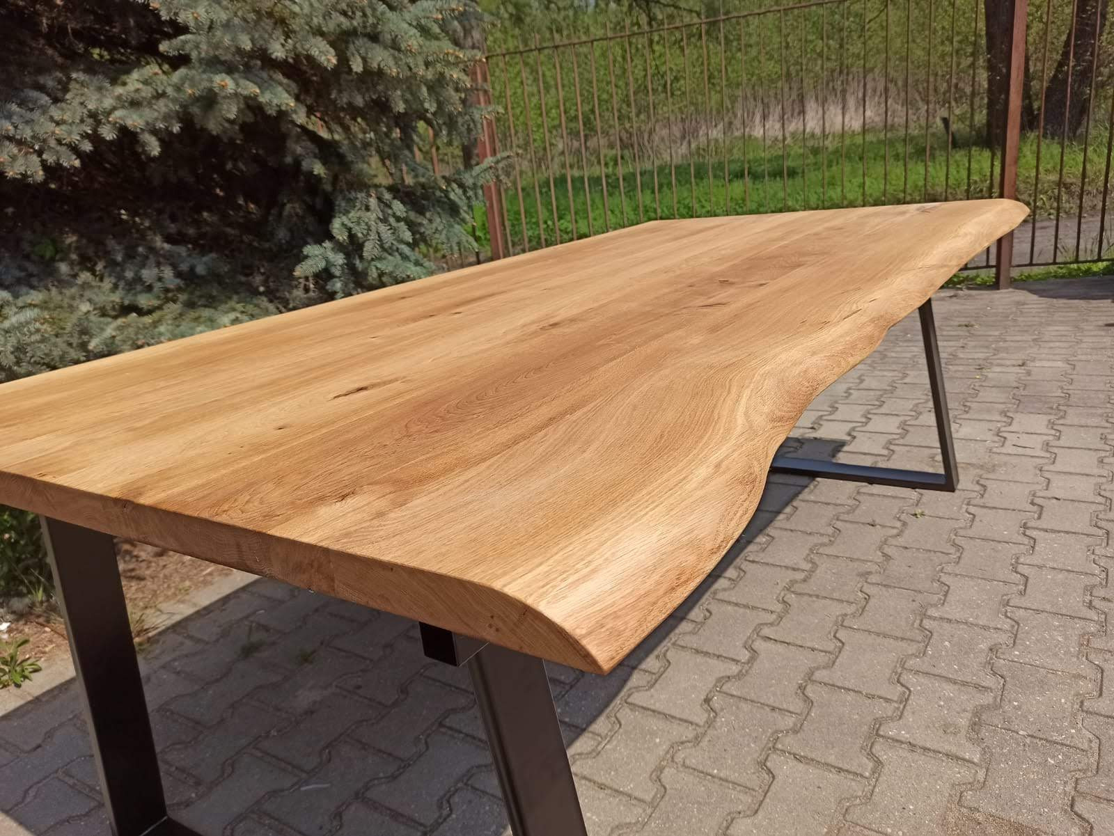 Surowy blat stołu z jasnego drewna o nieregularnych krawędziach, osadzony na czarnych, metalowych nogach w kształcie litery A, na tle kostki brukowej i zieleni.