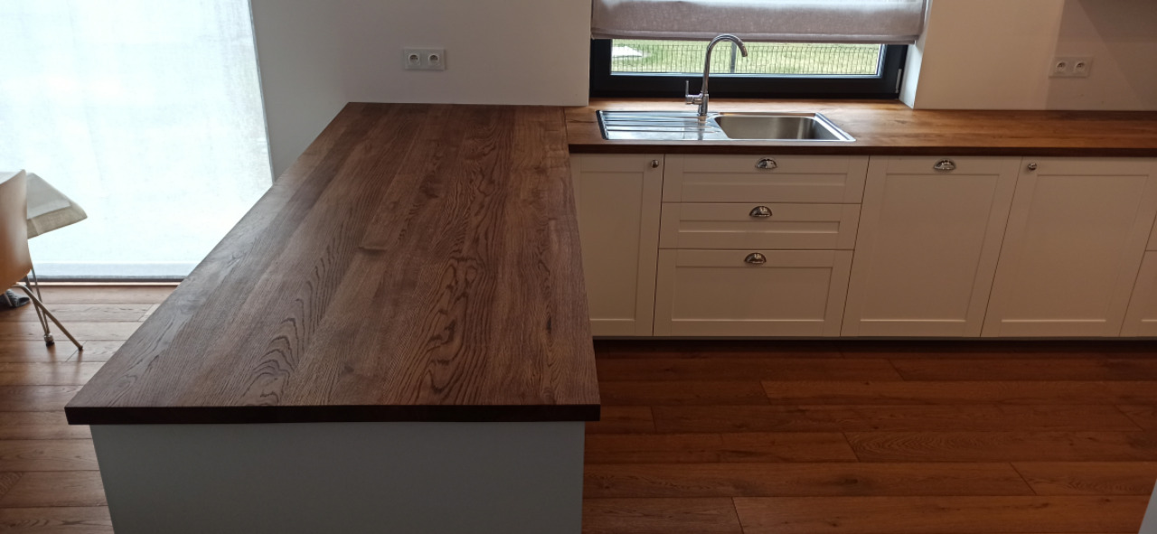 Kuchenny blat z ciemnego drewna o wyraźnym usłojeniu, zamontowany na białych szafkach w nowoczesnej kuchni, widok z boku na fragment blatu wyspowego.